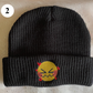 A black beanie labelled 2. It features a small, embroidered emoji face that has its eyes closed and many hearts floating around it.