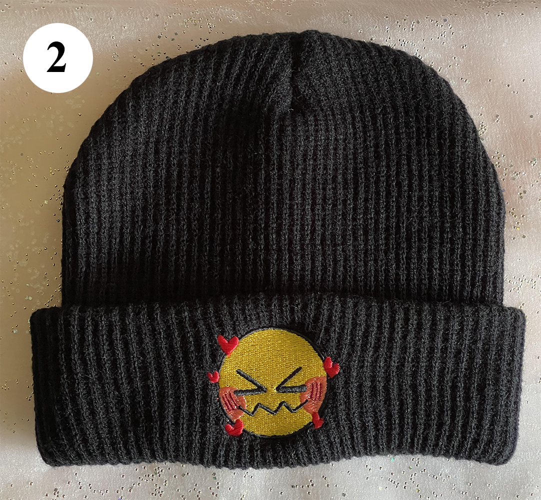 A black beanie labelled 2. It features a small, embroidered emoji face that has its eyes closed and many hearts floating around it.