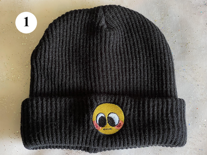 A black beanie labelled 1. It features a small, embroidered emoji face that is blushing and looking down.