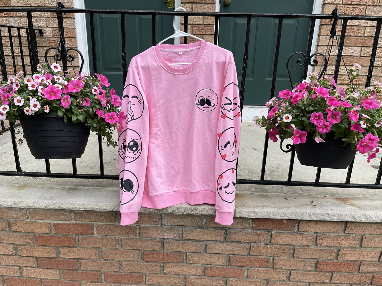 A pink Nice Emoji sweater hanging outside. It features one emoji design on the front, and three on each sleeve.