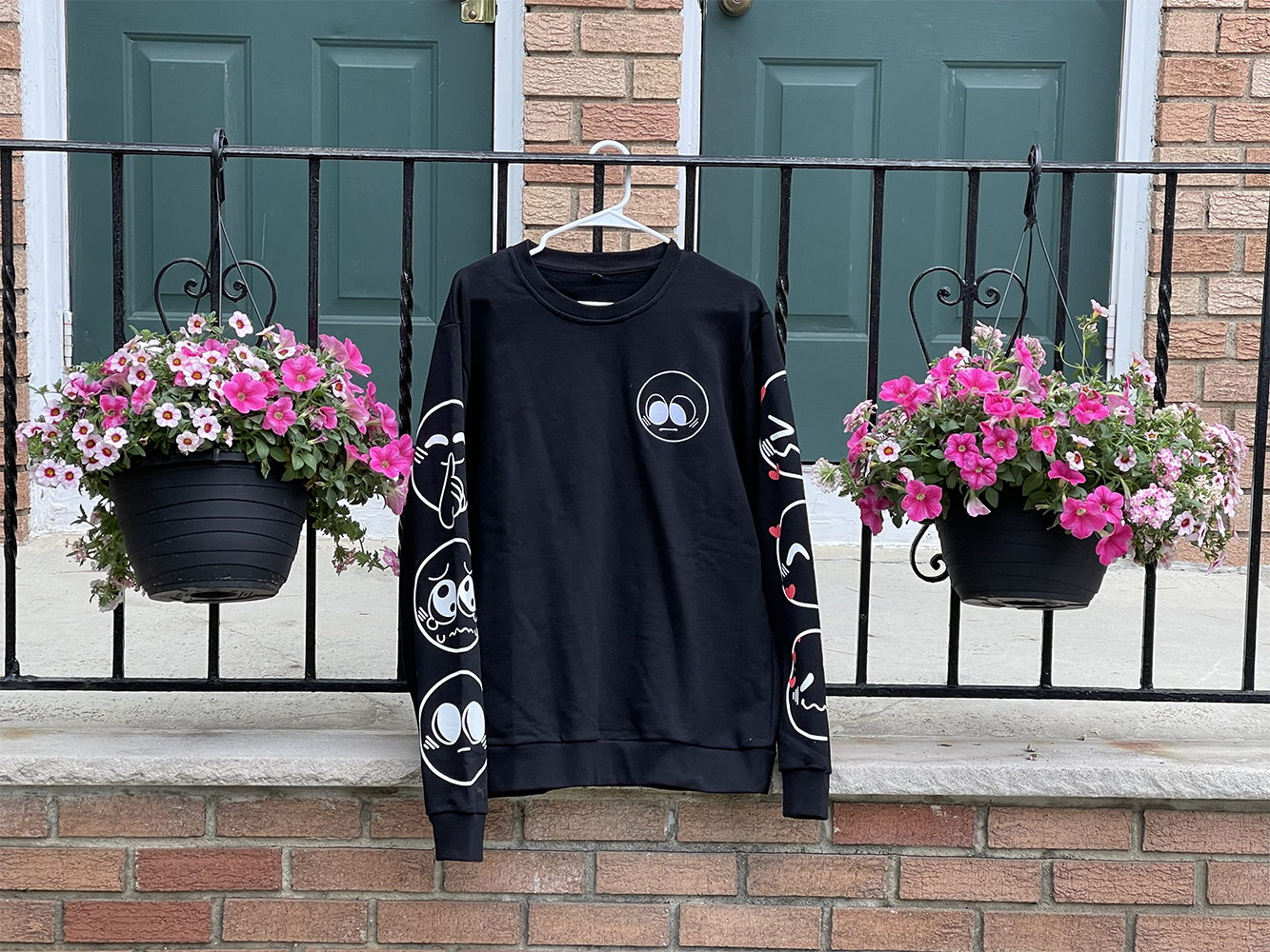 A black Nice Emoji sweater hanging outside. It features one emoji design on the front, and three on each sleeve.